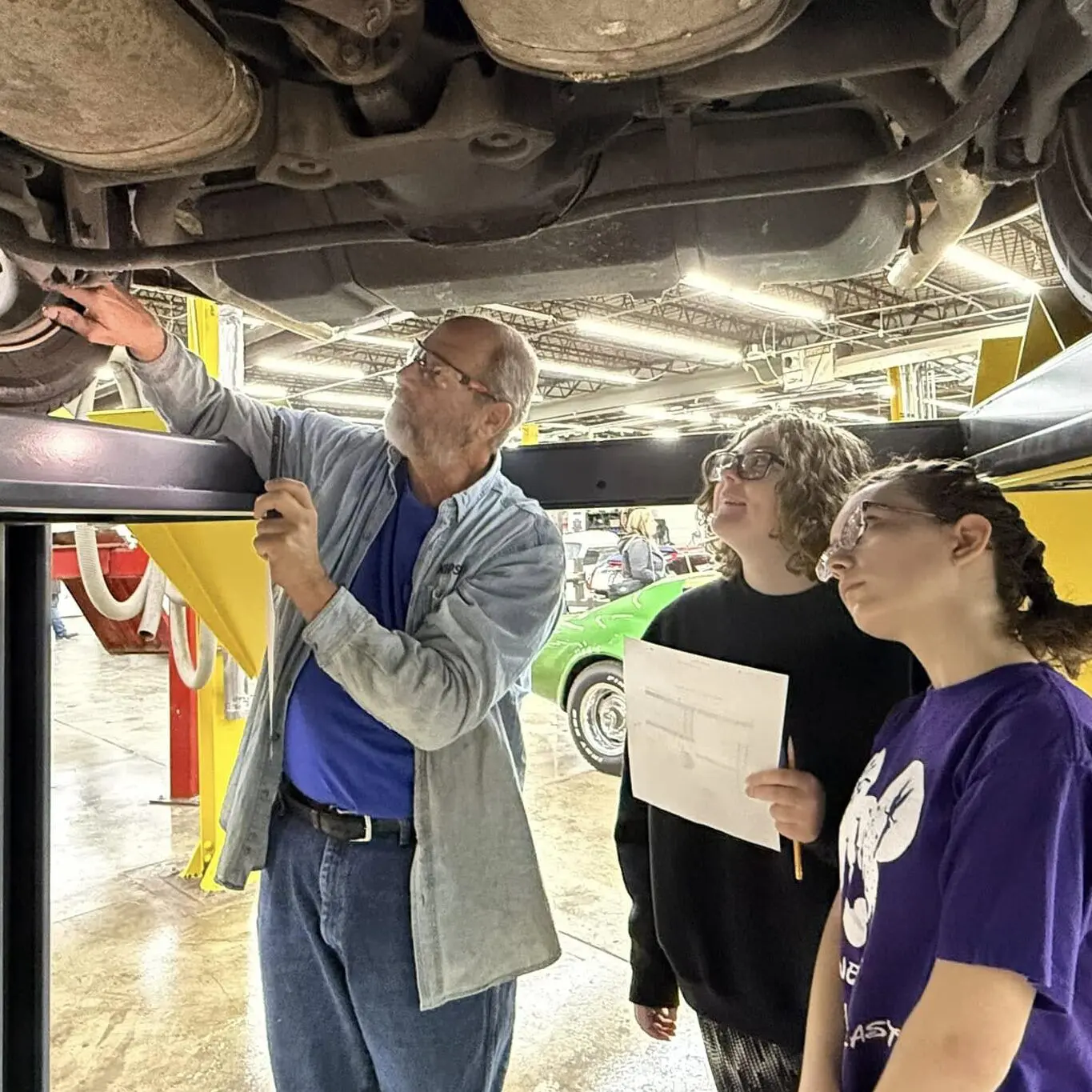 Two girls are shown the undercarriage of a vehicle on a lift